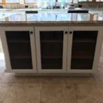 Kitchen island with cream-colored cabinetry, dark speckled granite countertop, three glass-door cabinets with dark interiors, and cork flooring