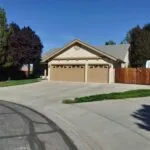 Single-story residential house with tan two-car garage, concrete driveway, and landscaped yard with evergreen trees under clear blue sky
