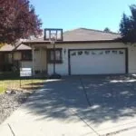 Single-story residential house with white exterior, two-car garage, driveway, and mature trees in front yard