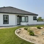 Modern white single-story house with black roof, dark-framed windows, landscaped yard with gravel bed and young shrubs