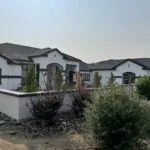 Modern white single-story home with dark roof, arched entry, landscaping with shrubs on arid lot