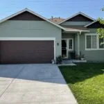 Mint green single-story house with dark brown garage door, concrete driveway, and manicured lawn under clear blue sky