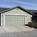 Single-story house with light gray siding, two white garage doors, dark roof, concrete driveway, and wooden fence.